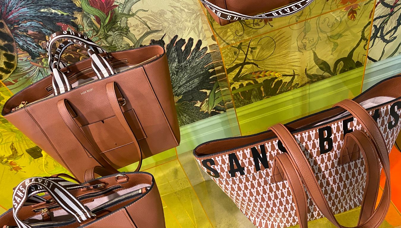 Three brown toned Sans Beast tote bags sit on yellow perspex plinths in front of a richly patterned wallpaper.