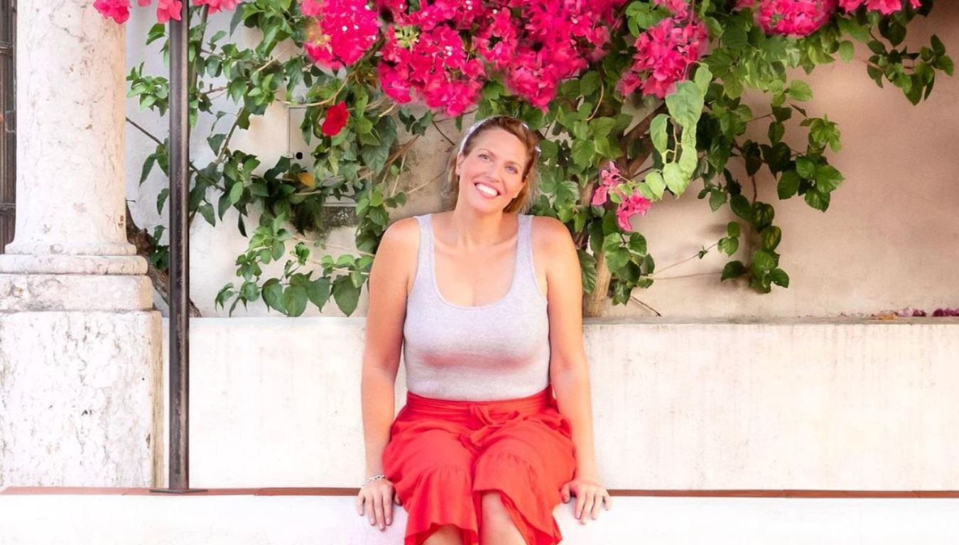 A woman wearing a red skirt sits under a brightly coloured bouganvillea plant, smiling broadly at the camera.