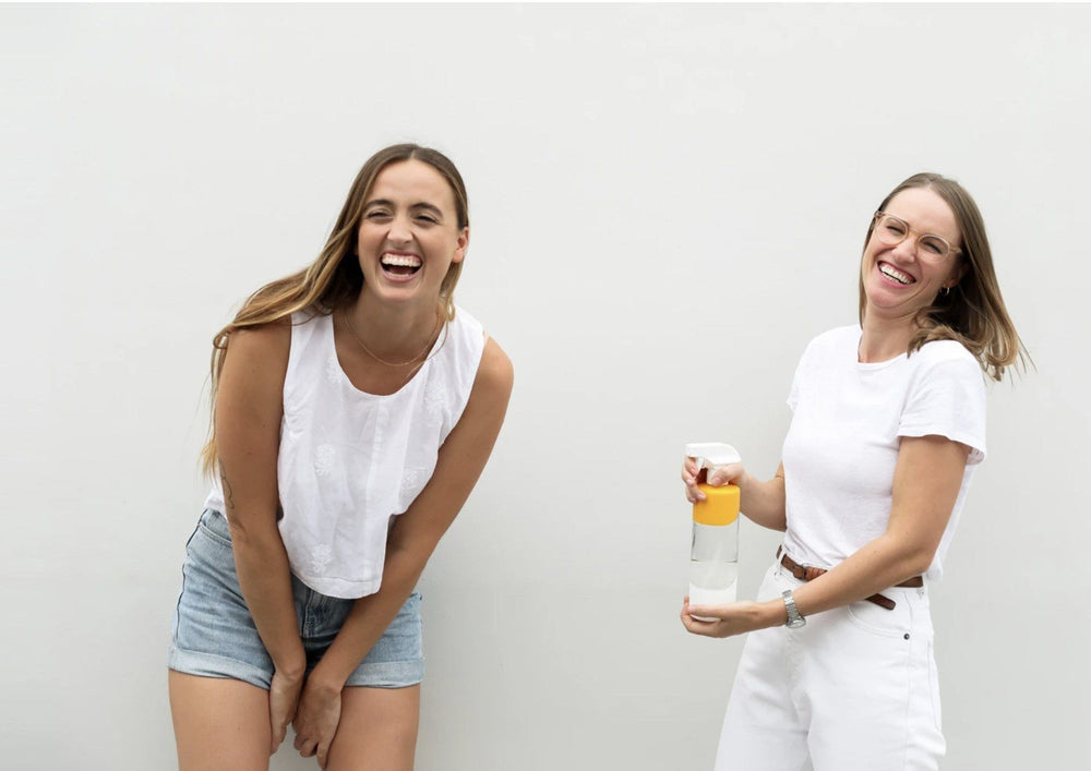 Image of Sian + Amy, founders of Pleasant State. Both women are laughing + standing in front of a light backdrop. Ami is holding a spray bottle containing the Pleasant State cleaning product in recyclable packaging.
