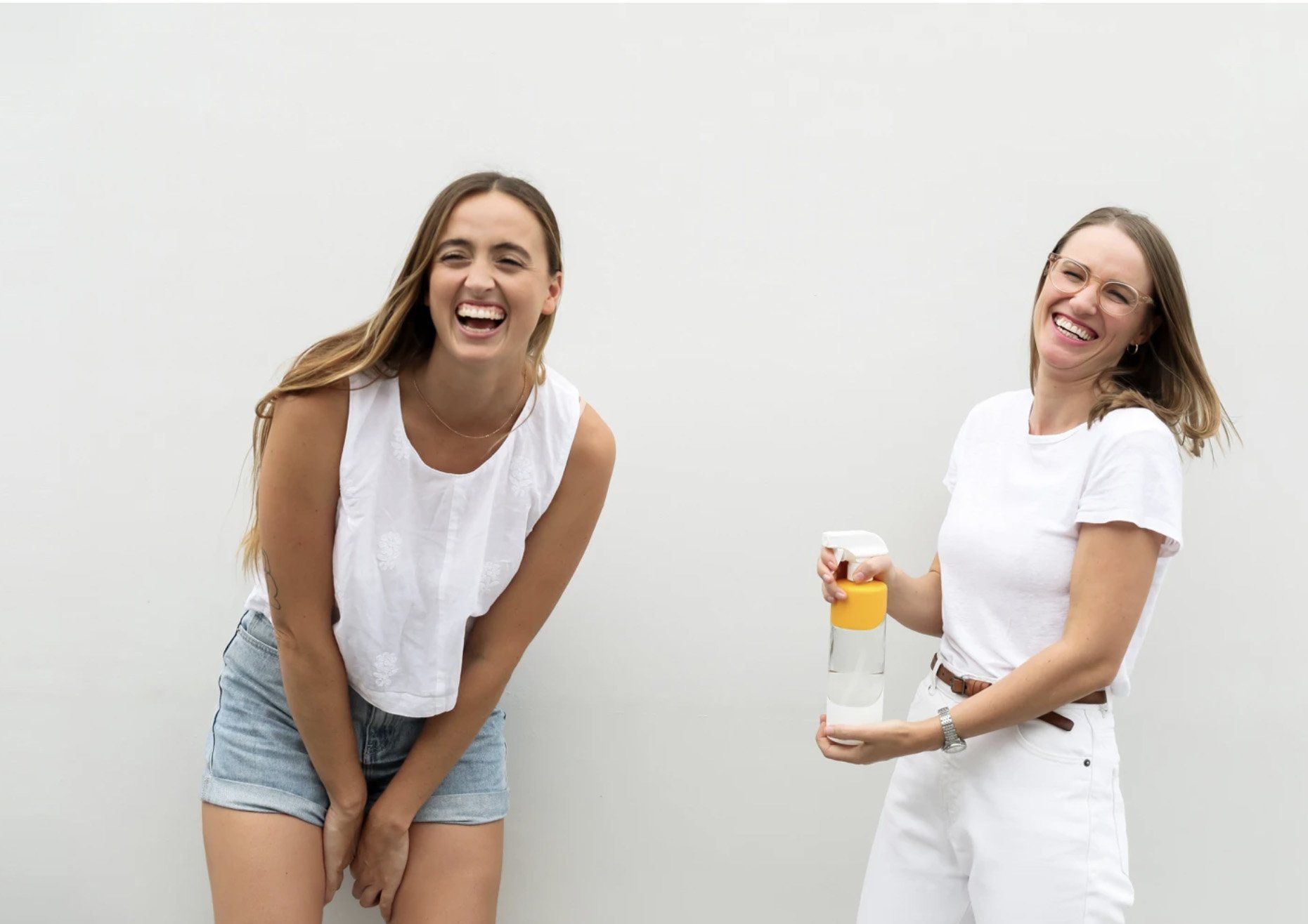 Image of Sian + Amy, founders of Pleasant State. Both women are laughing + standing in front of a light backdrop. Ami is holding a spray bottle containing the Pleasant State cleaning product in recyclable packaging.