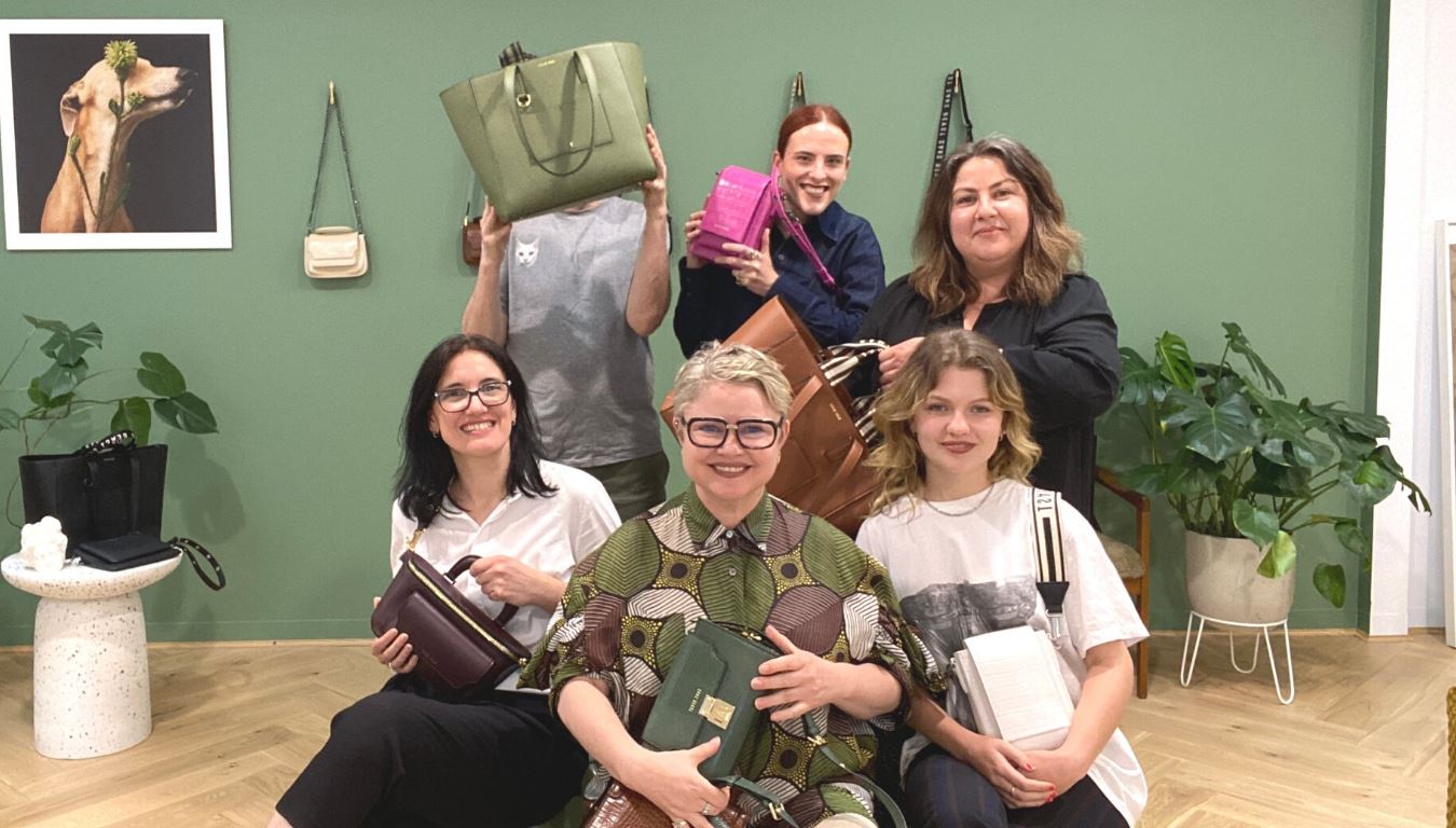 Image of five people sitting + standing in front of the camera holding up handbags.  In the background is a green painted wall, Sans Beast handbags hanging, a picture of a greyhound + some monstera plants