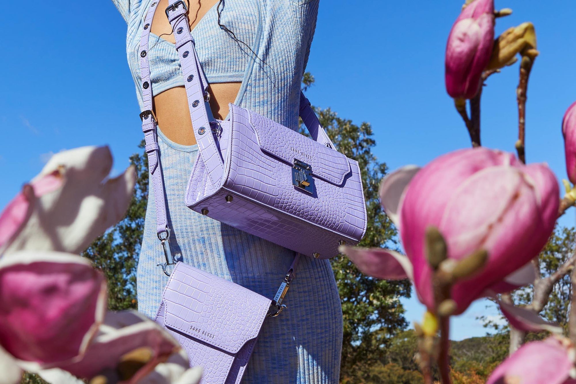 A woman wearing a light blue cutout dress and two lilac handbags reaching up to a bright blue sky.  She is surrounded by pink magnolia flowers.